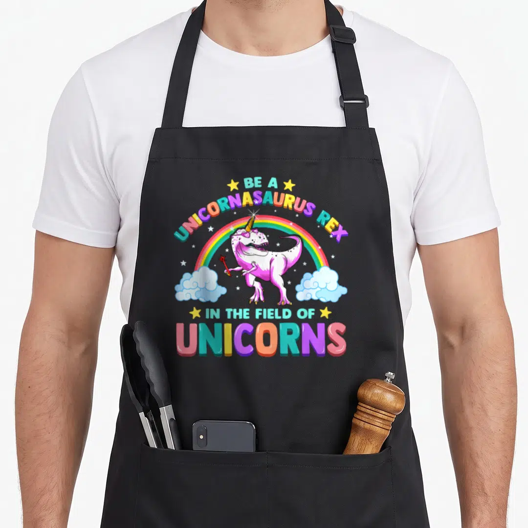 Unicornasaurus unicorn apron - Pink dinosaur holding wand under rainbow with stars and arched text — shown on a black cotton canvas apron against a white background