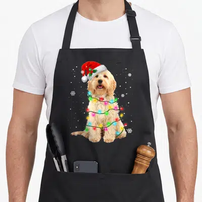 Tree goldendoodle apron - Golden dog wearing red santa hat wrapped in colorful lights with white snowflakes — shown on a black cotton canvas apron against a white background