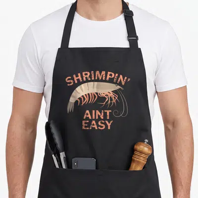 Shrimpin fishing aprons - Peach text SHRIMPIN AINT EASY over a grey and beige cartoon shrimp illustration — shown on a black cotton canvas apron against a white background