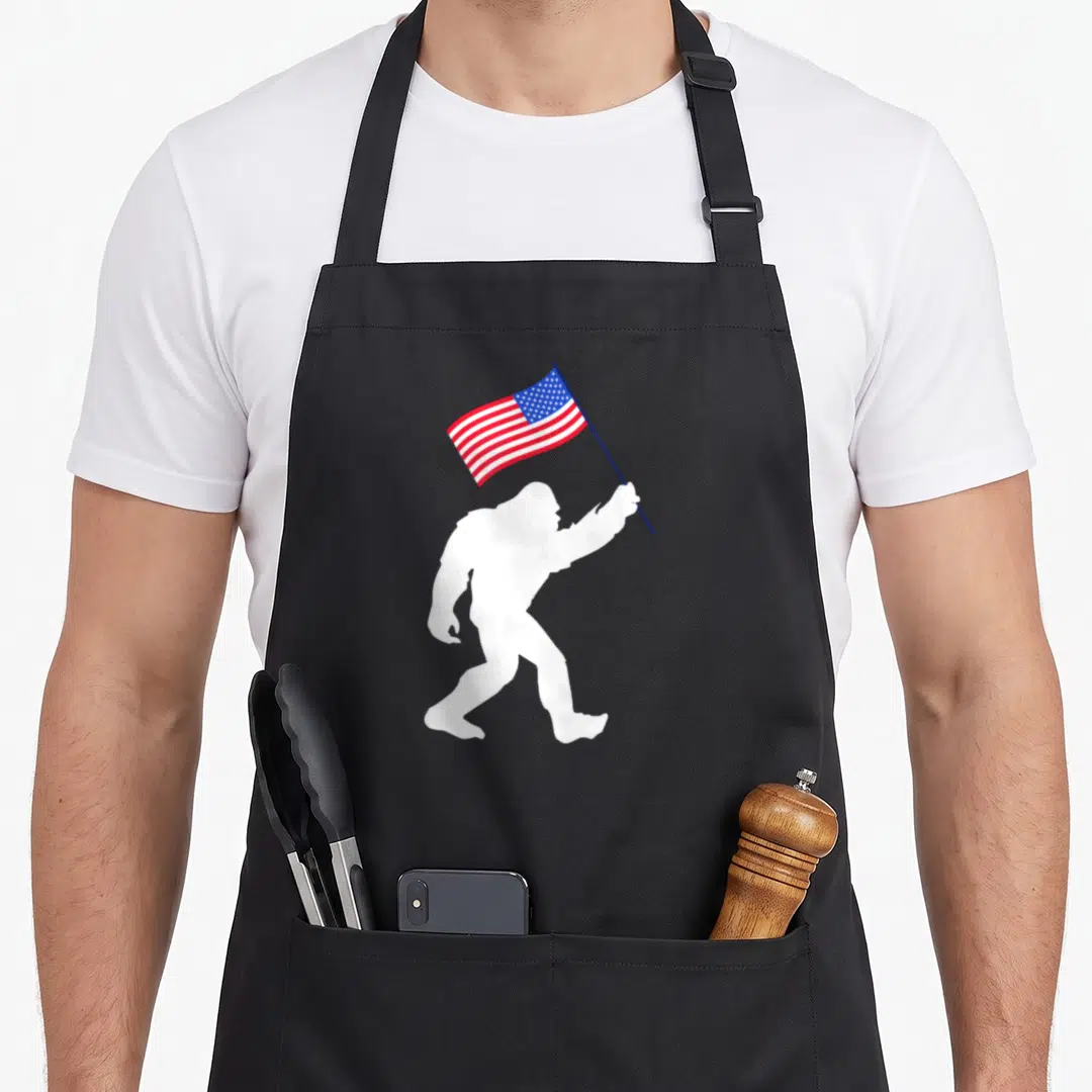 Cute bigfoot 4th of july apron - White bigfoot silhouette walking right holding waving red white and blue USA flag — shown on a black cotton canvas apron against a white background