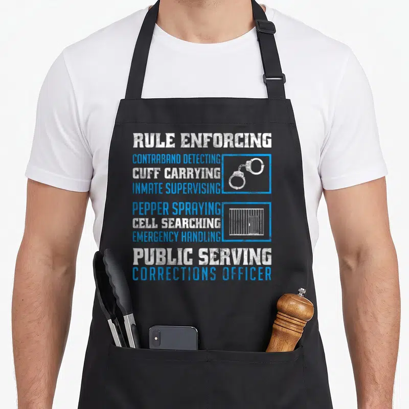 Correction retired officer apron - Stacked blue and white text listing corrections duties next to handcuff and jail bar — shown on a black cotton canvas apron against a white background