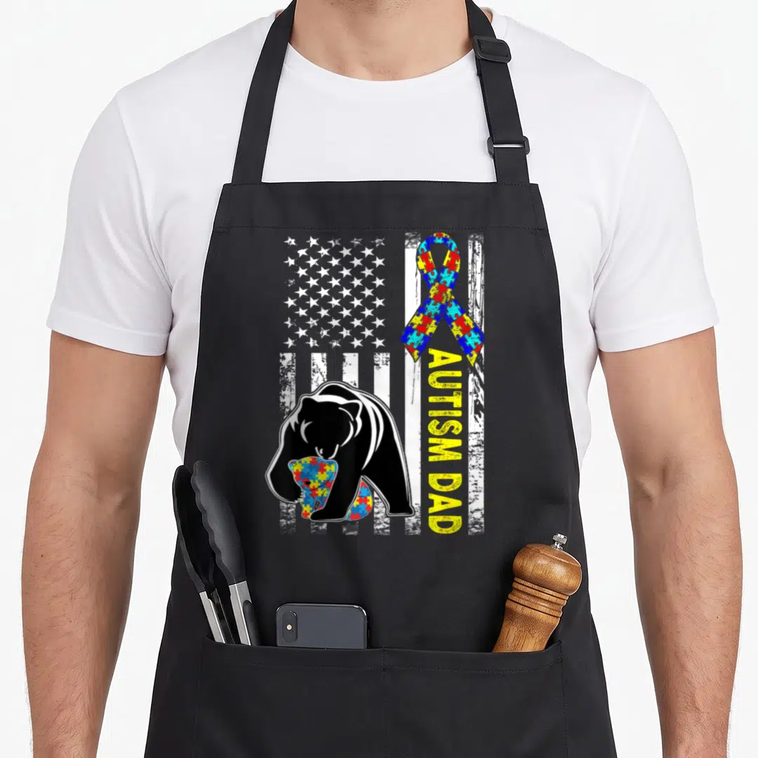 Autism father day apron - Black bear hugging puzzle cub on distressed US flag with vertical yellow text — shown on a black cotton canvas apron against a white background