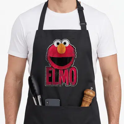 Smile elmo apron - Red cartoon character head above bold ELMO text in orange, white, and black — shown on a black cotton canvas apron against a white background