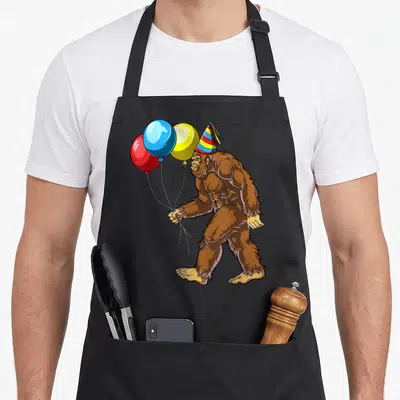 Birthday bigfoot apron - Cartoon brown bigfoot facing left wearing party hat and holding three balloons — shown on a black cotton canvas apron against a white background