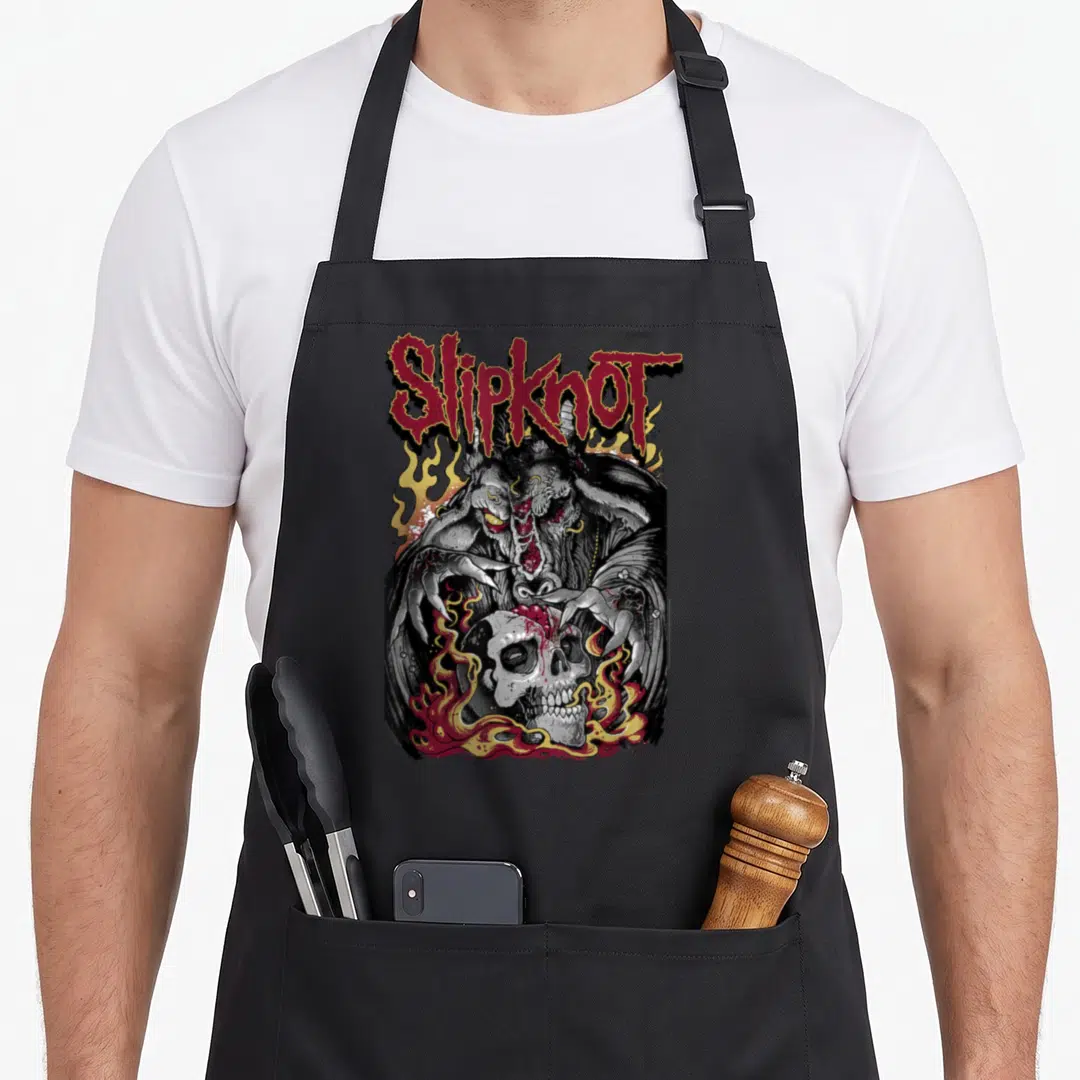 Slipknot heavy metal apron - Red arched text above a grey demon holding a white skull surrounded by yellow and red — shown on a black cotton canvas apron against a white background