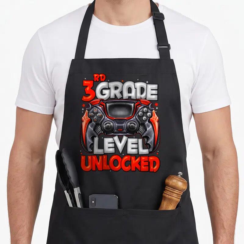 Unlocked game aprons - Red black and white game controller with RD 3 GRADE LEVEL UNLOCKED text and stars — shown on a black cotton canvas apron against a white background