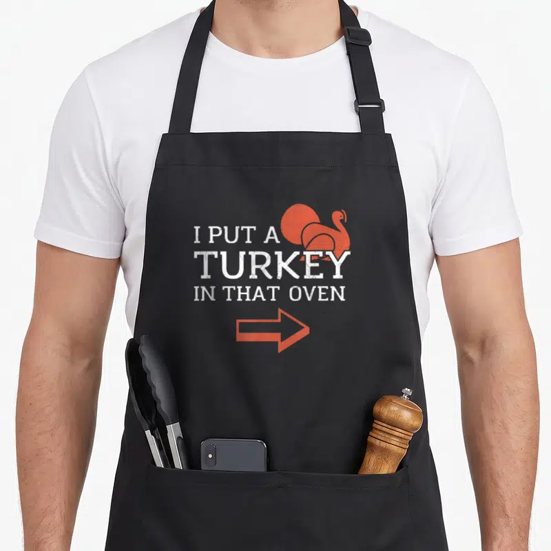 Oven turkey apron - White stacked text I PUT A TURKEY IN THAT OVEN with orange bird and arrow — shown on a black cotton canvas apron against a white background