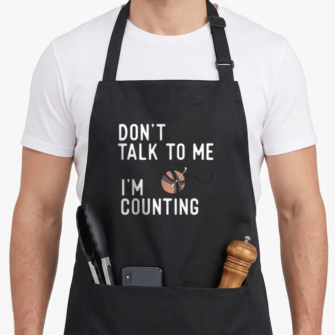 Talk crochet apron - Stacked white text reading Don't Talk to Me I'm Counting next to yarn ball — shown on a black cotton canvas apron against a white background