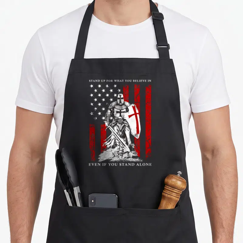 Knight american flag apron - Black and white knight holding a cross shield in front of a distressed red and white flag — shown on a black cotton canvas apron against a white background