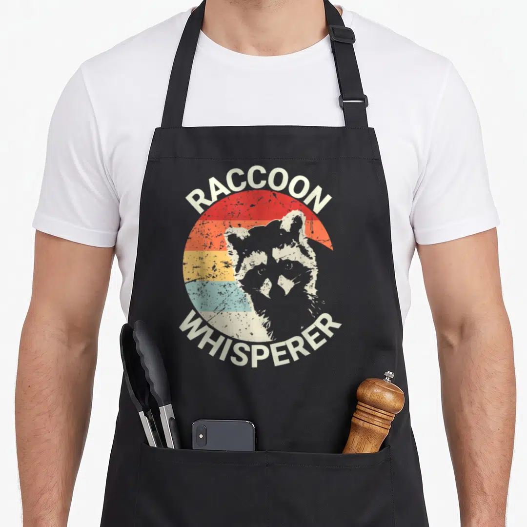 Raccoon pet apron - Raccoon Whisperer retro design with grey head on red orange yellow blue striped circle — shown on a black cotton canvas apron against a white background