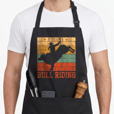 Bull riding western aprons - Grey bull rider silhouette over orange yellow green and red stripes with red text — shown on a black cotton canvas apron against a white background