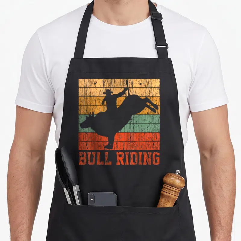 Bull riding western aprons - Grey bull rider silhouette over orange yellow green and red stripes with red text — shown on a black cotton canvas apron against a white background