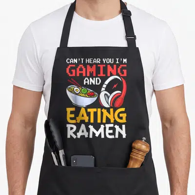 Eating gaming apron - Stacked text reading CAN'T HEAR YOU I'M GAMING AND EATING RAMEN above a cartoon ramen bowl — shown on a black cotton canvas apron against a white background