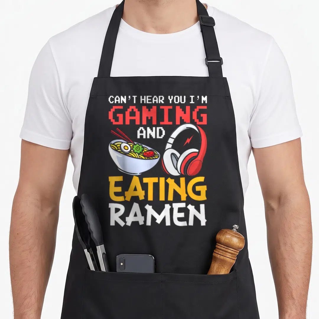 Eating gaming apron - Stacked text reading CAN'T HEAR YOU I'M GAMING AND EATING RAMEN above a cartoon ramen bowl — shown on a black cotton canvas apron against a white background