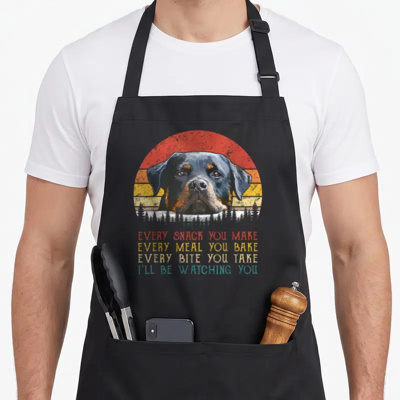 Snack rottie dog apron - Rottweiler head in front of red orange yellow striped sun with white forest silhouette — shown on a black cotton canvas apron against a white background