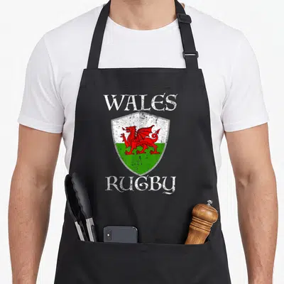 Rugby welsh apron - Distressed shield with red Welsh dragon on white and green background flanked by white text — shown on a black cotton canvas apron against a white background