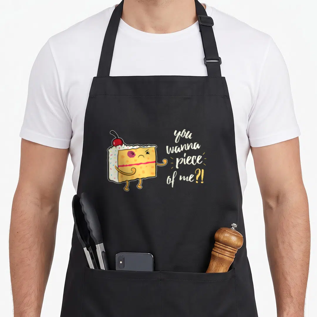 Funny cake baker apron - Cartoon cake slice with cherry and text you wanna piece of me in yellow white red — shown on a black cotton canvas apron against a white background