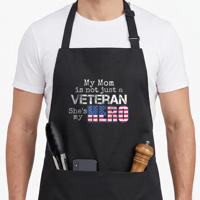 Veteran family aprons - Distressed white text reading My Mom Veteran Hero with flag pattern fill — shown on a black cotton canvas apron against a white background
