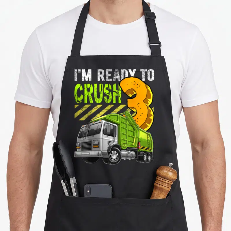 Garbage 3rd truck apron - Cartoon garbage truck with I'm ready to crush 3 text and hazard stripes — shown on a black cotton canvas apron against a white background
