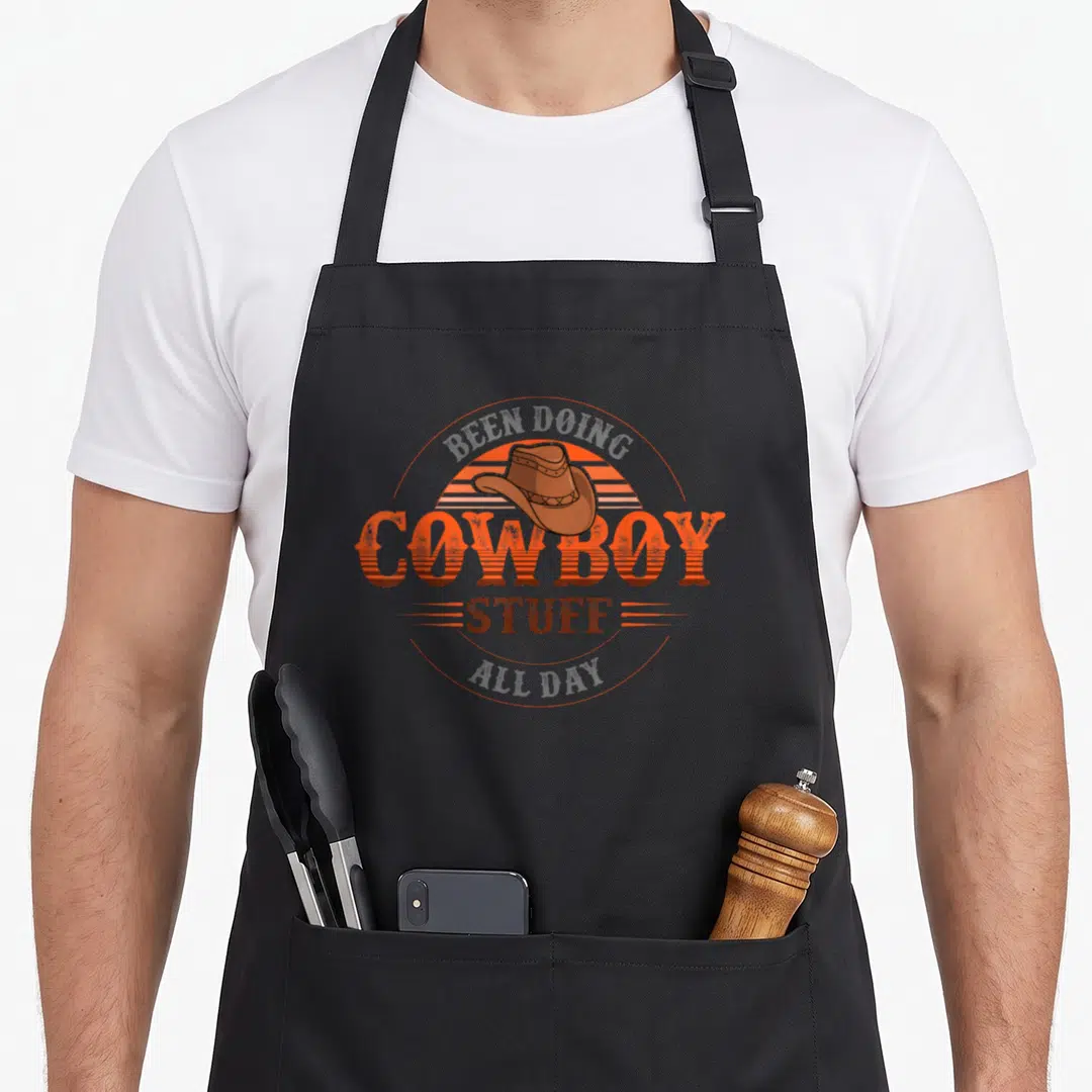 Doing cowboy apron - Retro brown cowboy hat over orange striped sun with BEEN DOING COWBOY STUFF ALL DAY text — shown on a black cotton canvas apron against a white background