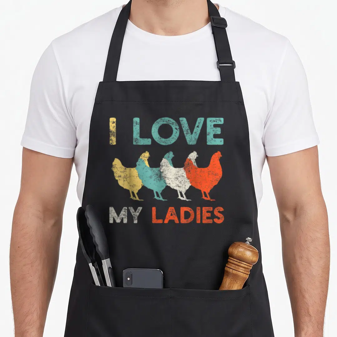 Farming chicken with apron - I Love My Ladies text with five colored hen silhouettes in yellow teal white and orange — shown on a black cotton canvas apron against a white background