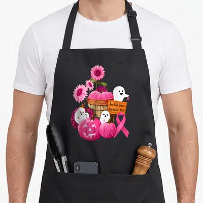 October pumpkin apron - Pink pumpkins, ghosts, sunflowers, wooden sign reading In October We Wear Pink, and large pink — shown on a black cotton canvas apron against a white background