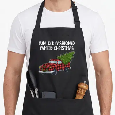 Family christmas tree apron - Red plaid truck carrying green tree with white text FUN OLD FASHIONED FAMILY CHRISTMAS — shown on a black cotton canvas apron against a white background