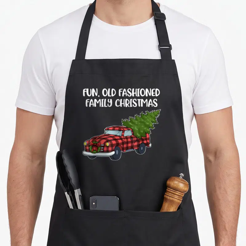 Family christmas tree apron - Red plaid truck carrying green tree with white text FUN OLD FASHIONED FAMILY CHRISTMAS — shown on a black cotton canvas apron against a white background