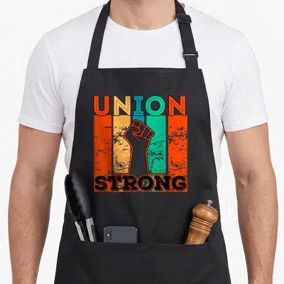 Funny strong union aprons - Raised fist between UNION STRONG text over orange teal yellow black vertical stripes retro — shown on a black cotton canvas apron against a white background