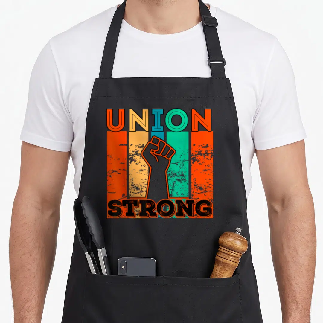 Funny strong union aprons - Raised fist between UNION STRONG text over orange teal yellow black vertical stripes retro — shown on a black cotton canvas apron against a white background