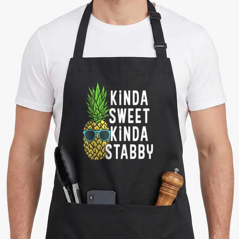 Kinda pineapple apron - Yellow cartoon pineapple with blue sunglasses beside stacked white text kinda sweet kinda stabby — shown on a black cotton canvas apron against a white background