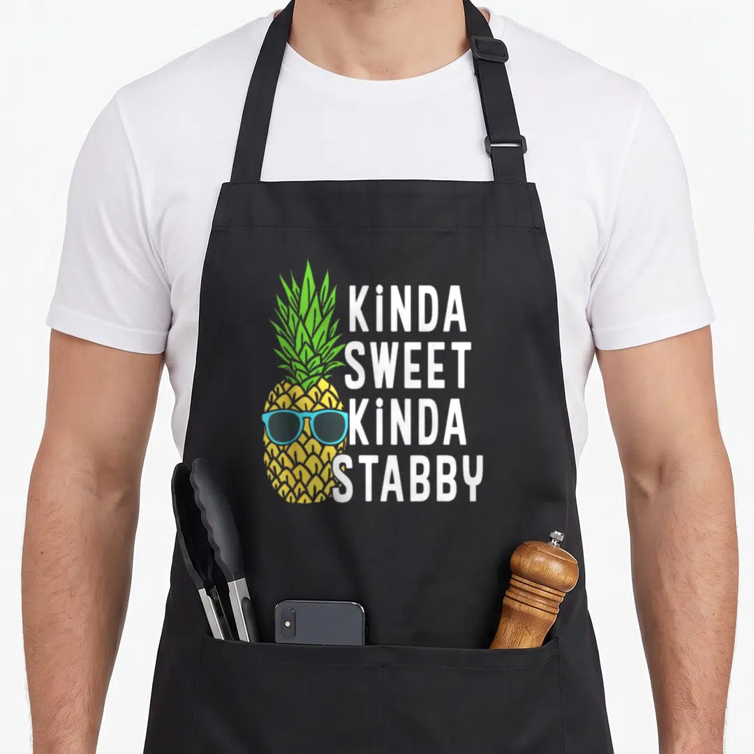 Kinda pineapple apron - Yellow cartoon pineapple with blue sunglasses beside stacked white text kinda sweet kinda stabby — shown on a black cotton canvas apron against a white background