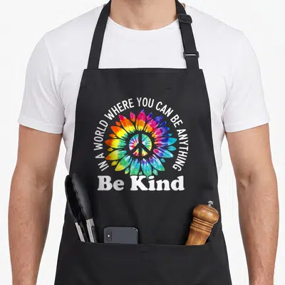 Anything karma apron - Tie-dye flower with peace sign and white text IN A WORLD WHERE YOU CAN BE ANYTHING, Be Kind — shown on a black cotton canvas apron against a white background