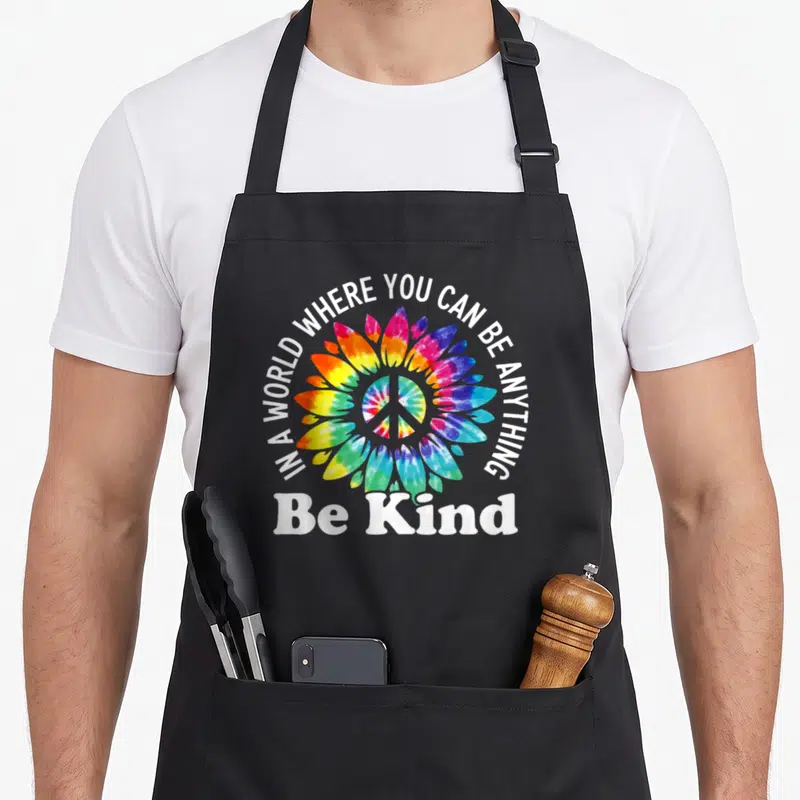 Anything karma apron - Tie-dye flower with peace sign and white text IN A WORLD WHERE YOU CAN BE ANYTHING, Be Kind — shown on a black cotton canvas apron against a white background