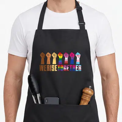 Rise pride apron - Seven raised fists in brown yellow purple blue pink above matching WE RISE TOGETHER text — shown on a black cotton canvas apron against a white background