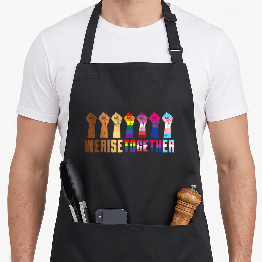 Rise pride apron - Seven raised fists in brown yellow purple blue pink above matching WE RISE TOGETHER text — shown on a black cotton canvas apron against a white background