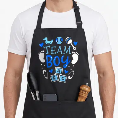 Gender baby apron - Team Boy text with blue duck, ABC blocks, rattle, hearts, footprints, and safety pins in circular — shown on a black cotton canvas apron against a white background