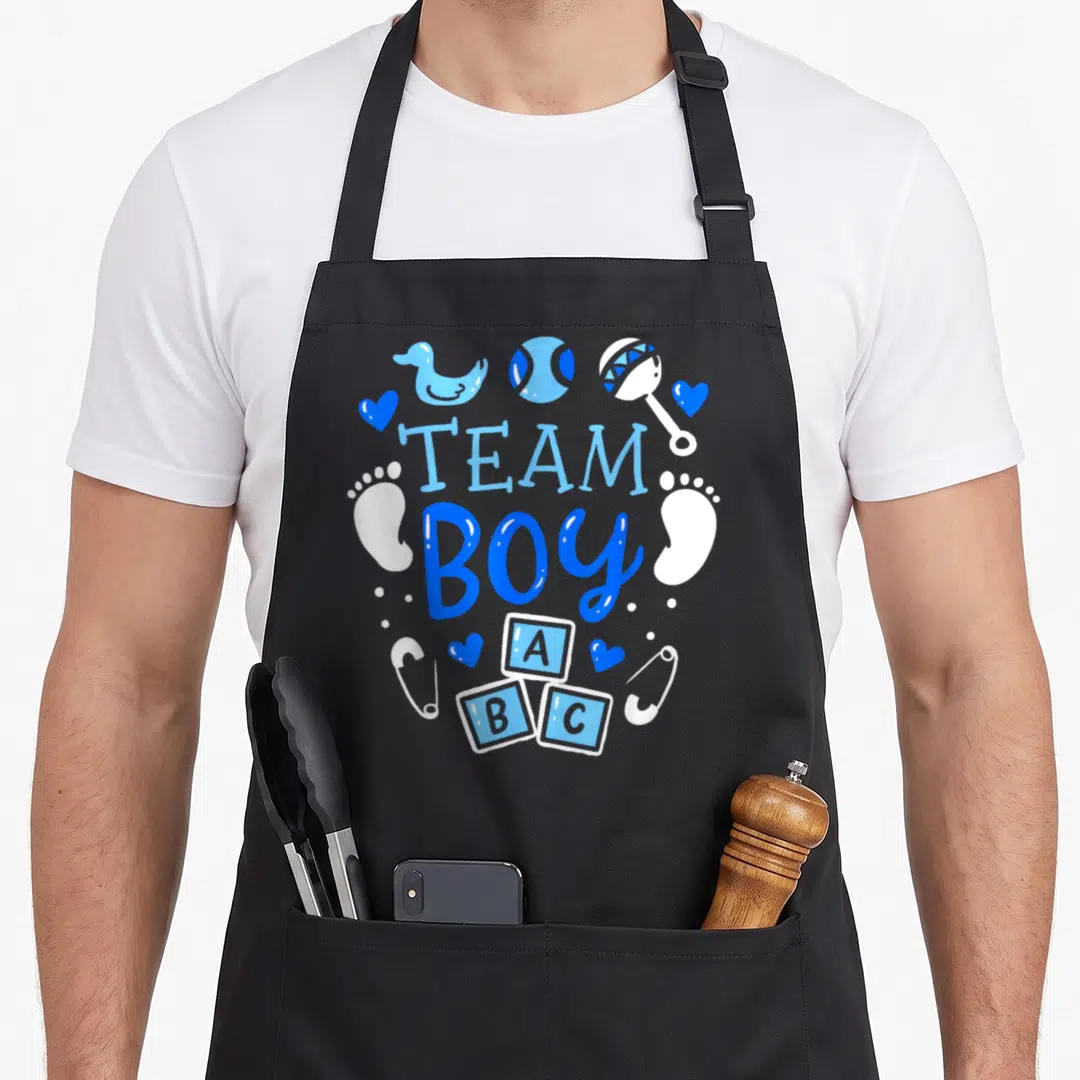 Gender baby apron - Team Boy text with blue duck, ABC blocks, rattle, hearts, footprints, and safety pins in circular — shown on a black cotton canvas apron against a white background