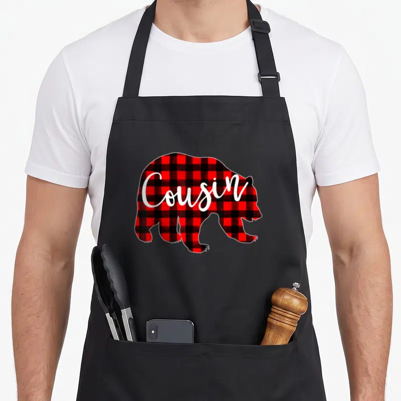 Cousin buffalo plaid christmas apron - Bear silhouette filled with red black plaid pattern and white Cousin script text — shown on a black cotton canvas apron against a white background