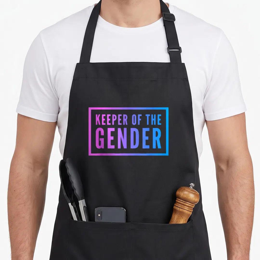 Keeper surprise party apron - KEEPER OF THE GENDER text in pink to blue gradient inside a rectangular border — shown on a black cotton canvas apron against a white background