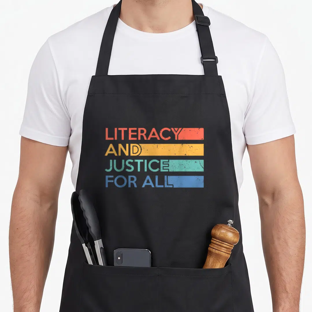 Literacy librarian apron - Stacked text LITERACY AND JUSTICE FOR ALL with red yellow green blue horizontal bars — shown on a black cotton canvas apron against a white background