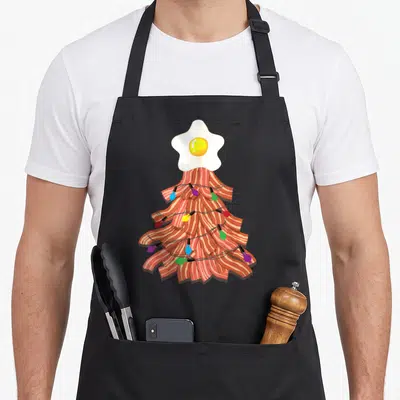 Christmas bacon apron - Cartoon bacon tree with fried egg top and multicolored string lights — shown on a black cotton canvas apron against a white background