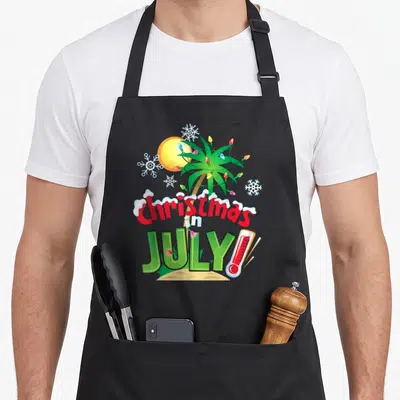 July christmas vacation apron - Palm tree with lights, snowflakes, sun, and thermometer on sand — shown on a black cotton canvas apron against a white background