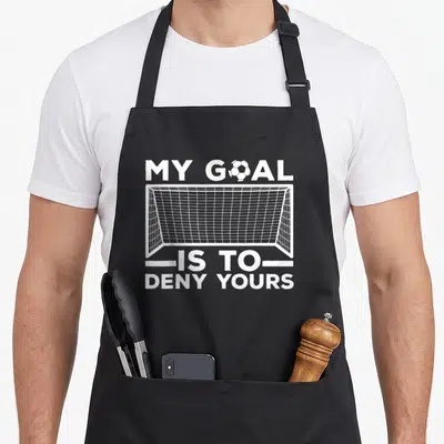 Defender funny soccer apron - White soccer goal graphic with text My Goal Is To Deny Yours and a ball replacing — shown on a black cotton canvas apron against a white background