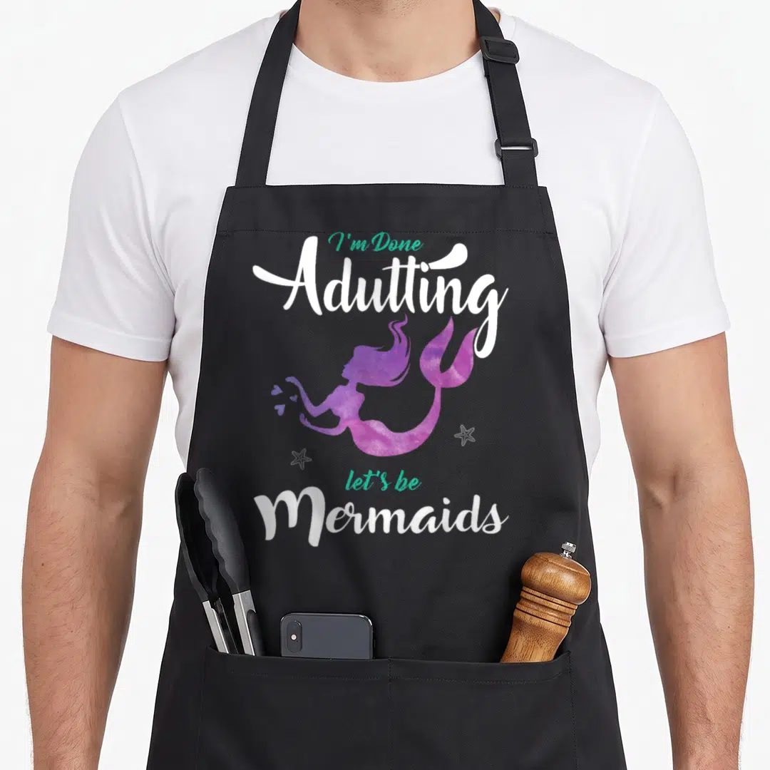 Adulting mermaid apron - Purple watercolor mermaid between white and teal text with two starfish on black background — shown on a black cotton canvas apron against a white background