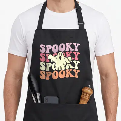Spooky ghost halloween apron - Stacked SPOOKY text in purple pink cream orange with cream cartoon ghost overlay — shown on a black cotton canvas apron against a white background