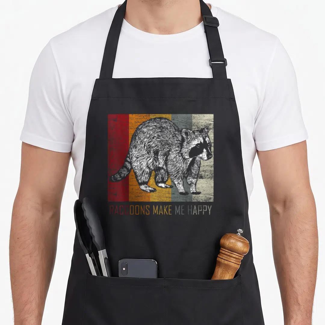 Racoon four raccoon apron - Grey raccoon illustration over six colored stripes with matching rainbow text below — shown on a black cotton canvas apron against a white background