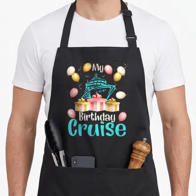 Festive birthday apron - Cruise ship illustration with balloons and three gift boxes under My Birthday Cruise text — shown on a black cotton canvas apron against a white background