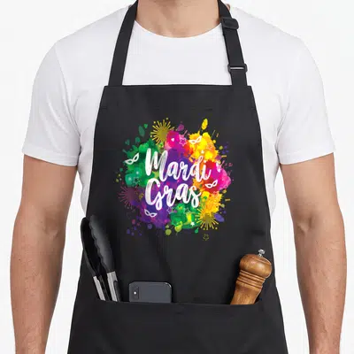 Mardi fat chef apron - White script text over purple green yellow white and pink watercolor splatters with masks — shown on a black cotton canvas apron against a white background
