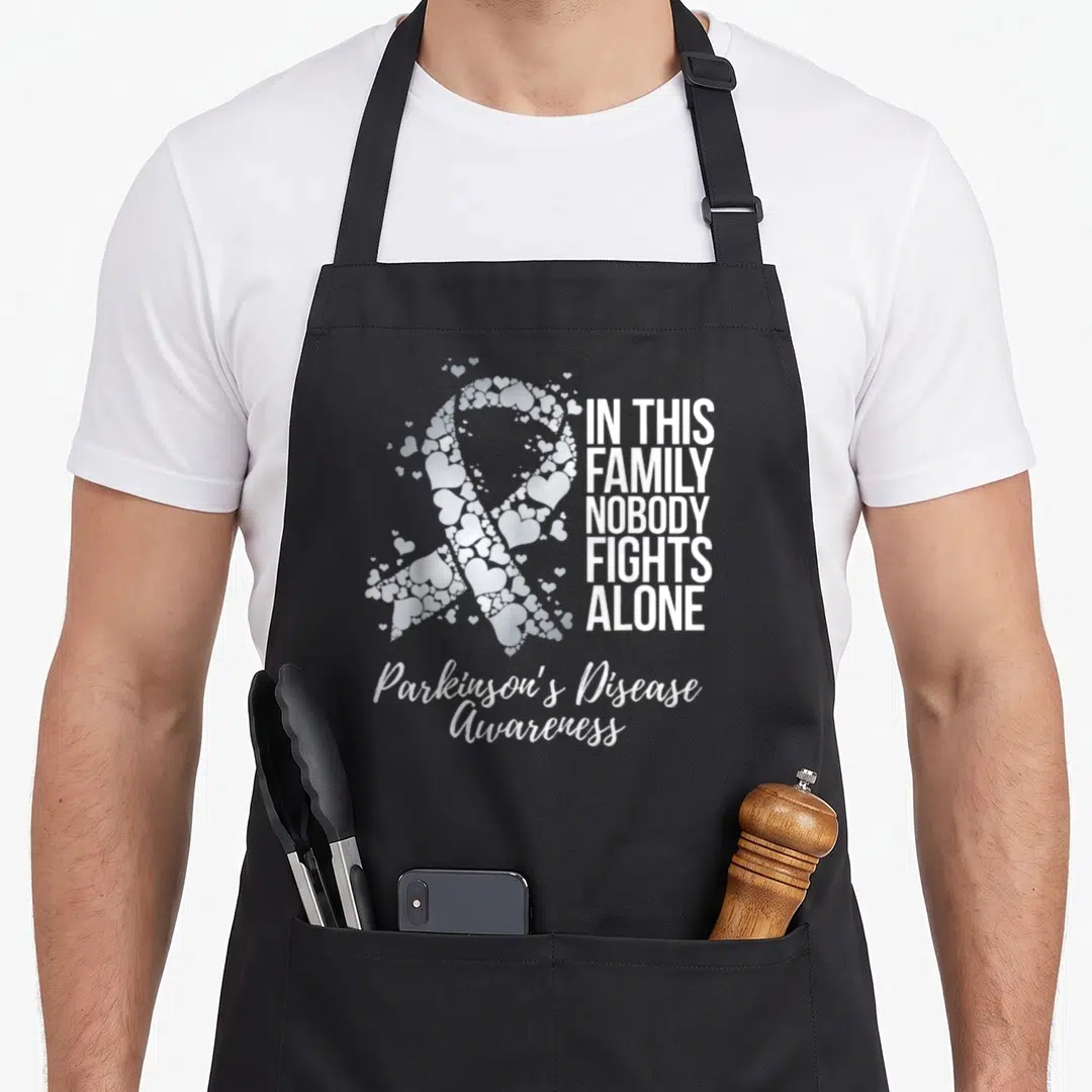 Support parkinson family aprons - White awareness ribbon made of small hearts next to block text IN THIS FAMILY NOBODY — shown on a black cotton canvas apron against a white background